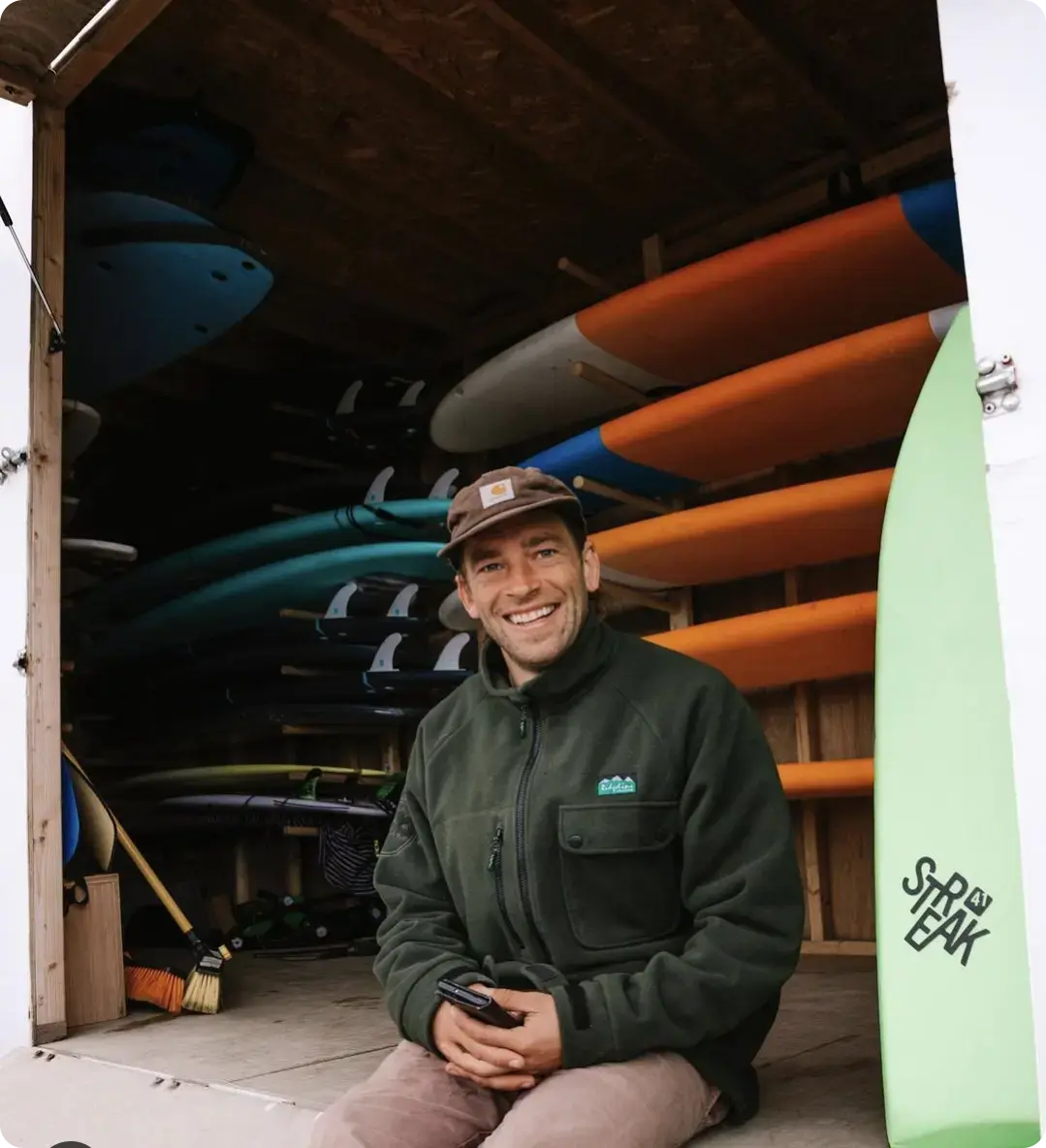 Tom Sherrin, the founder of Tom's Surf Hire, sitting in front of rental surfboards.